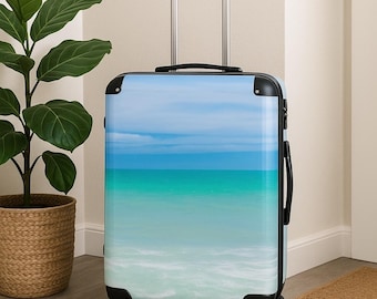 May include: A rolling suitcase with a beach scene design. The image on the suitcase features a blue sky, turquoise water, and white waves. The suitcase has a black handle, black trim, and four black wheels.