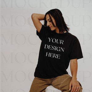 May include: A black t-shirt with the text "YOUR DESIGN HERE" printed on the front. The t-shirt is being worn by a person sitting on a white stool. The person is wearing brown trousers and white trainers.