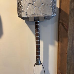 May include: A silver and gray hammer with a brown handle and a leather loop. The hammer head has a cracked texture and decorative detailing. The handle is wrapped with brown leather and silver bands.