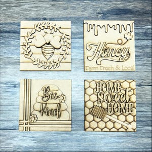 May include: Four wooden signs with bee-themed designs. The first sign features a bee in a wreath, the second sign says "Honey Farm Fresh & Local", the third sign says "Bee Kind" with a beehive, and the fourth sign says "Home Sweet Home" with a honeycomb pattern.