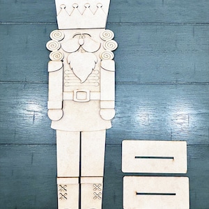 May include: Wooden nutcracker cutout with a crown, mustache, and beard. The cutout is unpainted and ready to be decorated. Two rectangular wooden pieces are included for adding a personalized message.