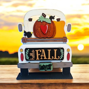 May include: White wooden truck with a red heart pumpkin, two gourds, and two black birds in the bed. The truck has a black sign on the back that says "hello FALL" with green leaves on either side. The truck has a license plate that says "HARVEST".