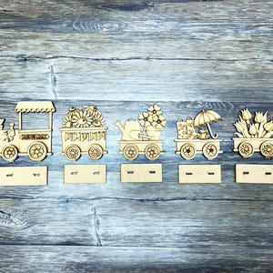 May include: Wooden train engine and cars decorated with spring flowers, a watering can, rain boots, and an umbrella. The train cars have the words "Springtime Charms" printed on them.