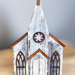 May include: A white wooden church model with a brown cross on top and brown trim around the windows and door. The church has a whitewashed, distressed look.