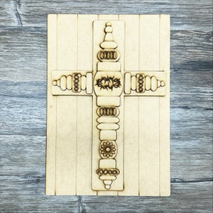 May include: A wooden cross with a decorative design featuring a crown of thorns, beads, and a flower. The cross is on a light brown background.