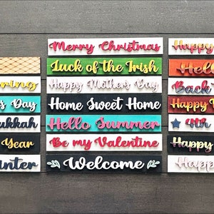 May include: A collection of 16 wooden signs with various holiday and seasonal greetings. The signs are painted in different colors and feature a variety of fonts. The signs include greetings such as "Merry Christmas", "Happy Birthday", "Hello Spring", "Happy Father's Day", "Happy Hanukkah", "Happy New Year", "Hello Winter", "Luck of the Irish", "Happy Mother's Day", "Home Sweet Home", "Hello Summer", "Be my Valentine", "Welcome", "Happy Halloween", "Happy Easter", "Back to School", "Happy Thanksgiving", "4th of July", and "Hello Fall".