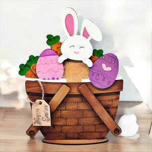 May include: Wooden Easter basket decoration featuring a white bunny with pink ears, carrots, and decorated eggs. The basket has a handle and a tag that says "Happy Easter". The eggs are pink and purple. The basket is brown.