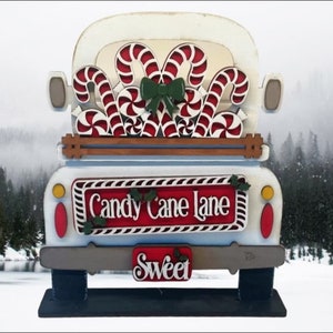 May include: White wooden truck with a red and white candy cane design. The truck is carrying a stack of candy canes and has a sign that says "Candy Cane Lane" and "Sweet".