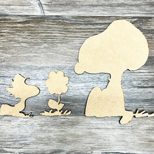 May include: Wooden cutouts of Snoopy, Woodstock, and a flower. The Snoopy cutout is sitting down, looking at the flower. The Woodstock cutout is lying down, looking at the flower. The flower is in the middle, with leaves and grass.