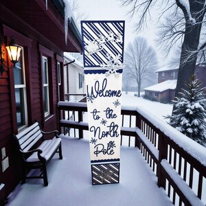 May include: A tall, rectangular wooden sign with the words "Welcome to the North Pole" in navy blue script. The sign features snowflake and Santa Claus cutouts, and is decorated with navy blue and white striped patterns. The sign is placed on a snow-covered porch.