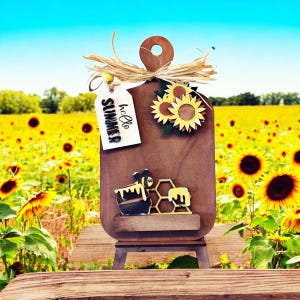 May include: A wooden decorative cutting board with a sunflower and bee theme. The board has a handle and a small shelf. It features a tag that says "hello SUMMER". The background is a field of sunflowers under a blue sky.