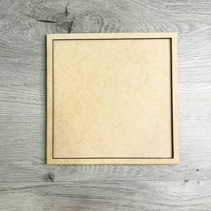 May include: A square, unfinished wooden sign with a thin black border. The sign is made of light brown wood and sits on a gray wooden surface. The sign is blank and ready for customization.