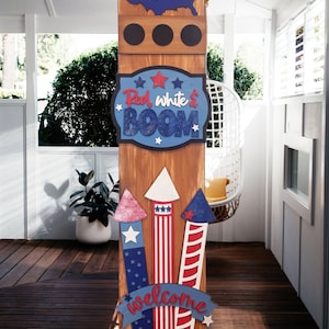 May include: Wooden sign with a red, white, and blue patriotic theme. It features a heart-shaped US map, a sign that says "Red White & Boom", and fireworks with a welcome banner.