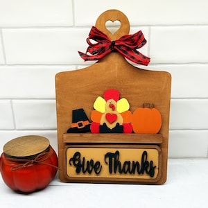 May include: Wooden cutting board with a shelf and a sign that says "Give Thanks". The board is decorated with a turkey, a pumpkin, and a pilgrim hat.