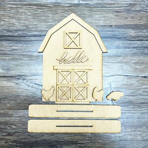 May include: Wooden cutout of a barn with a sign that says "hello" and two chickens. The barn has a window and a door with a crosshatch design. The barn is on a platform with two shelves.