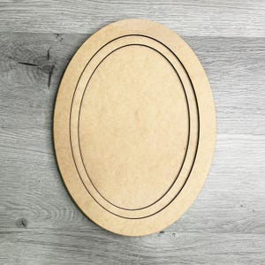 May include: A light brown, oval-shaped wooden plaque with a recessed oval design. The plaque has a smooth surface and is suitable for crafts or home decor projects. The design features two concentric oval outlines, creating a layered effect.