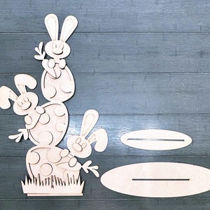May include: Wooden Easter bunny craft kit with three bunnies and eggs. The kit includes two oval-shaped bases with slots for holding the bunnies and eggs.