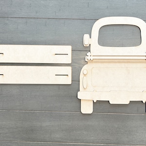 May include: A wooden cutout of a vintage pickup truck with a blank space in the back. The truck is light brown and has a white background. There are two wooden planks with slots on either side.