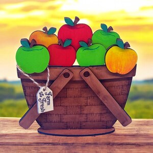 May include: A wooden basket filled with colorful wooden apples. The basket has a brown wooden handle and a sign that reads "Farm Fresh".