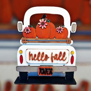 May include: White wooden truck with a bed full of orange pumpkins and a black bird on top. The truck has a sign on the back that says "hello fall" and a license plate that says "pumpkins".