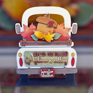 May include: A white wooden truck with a bed full of fall leaves and an acorn. The truck has a sign that reads "Leaves are falling, Autumn is calling, Sweater Weather".
