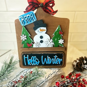 May include: Wooden cutting board with a snowman, two pine trees, and a red cardinal. The board has a red and black plaid bow and the text "BRRR" and "Hello Winter".