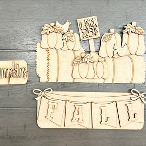 May include: Wooden craft kit for making a fall-themed pumpkin patch scene. The kit includes a sign that says "U-Pick 25¢", a banner that says "FALL", and various pumpkin shapes.