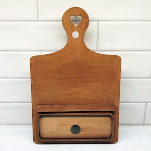 May include: A wooden cutting board with a heart-shaped cutout in the handle and a small drawer underneath. The board is brown and has a natural wood finish.