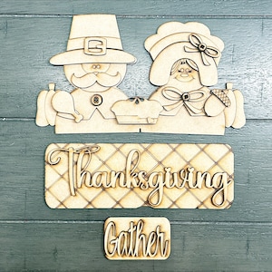 May include: Wooden cutout Thanksgiving decorations featuring a pilgrim man and woman with a banner that reads "Thanksgiving Gather".