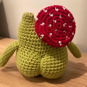 Op de afbeelding: Een gehaakt knuffeldier in de vorm van een paddenstoel. De paddenstoel is groen met een rood-witte gehaakte dop.