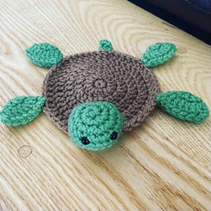 May include: A crocheted turtle coaster with a brown shell and green limbs. The turtle has black eyes and a green head.