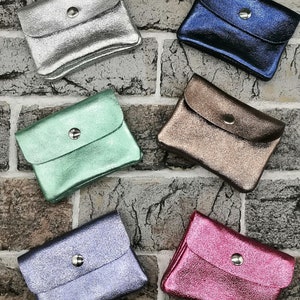 May include: A collection of small, metallic coin purses in various colors, including silver, blue, bronze, green, purple, and pink. Each purse has a flap closure secured by a silver snap. The purses are arranged on a brick-patterned surface.
