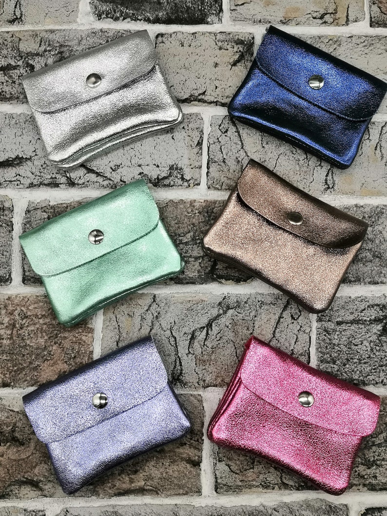 May include: A collection of small, metallic coin purses in various colors, including silver, blue, bronze, green, purple, and pink. Each purse has a flap closure secured by a silver snap. The purses are arranged on a brick-patterned surface.