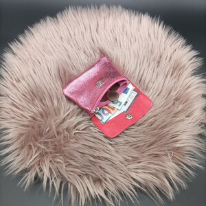 May include: A small, open pink leather coin purse with a metallic sheen, containing coins and paper currency. The interior is red, and the purse is secured with a snap closure. The purse is resting on a fluffy, light pink surface.