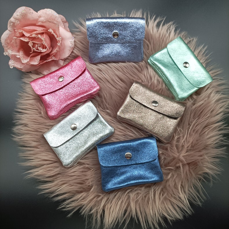May include: A collection of small, glittery coin purses in various colors, including pink, silver, blue, and gold. Each purse has a flap closure with a silver snap. The purses are arranged on a fluffy, pink surface, with a decorative pink flower in the background.