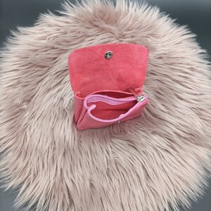 May include: A small, open, pink leather coin purse with a matching pink zipper pouch inside. The purse has a snap closure and is set against a fluffy, light pink background.