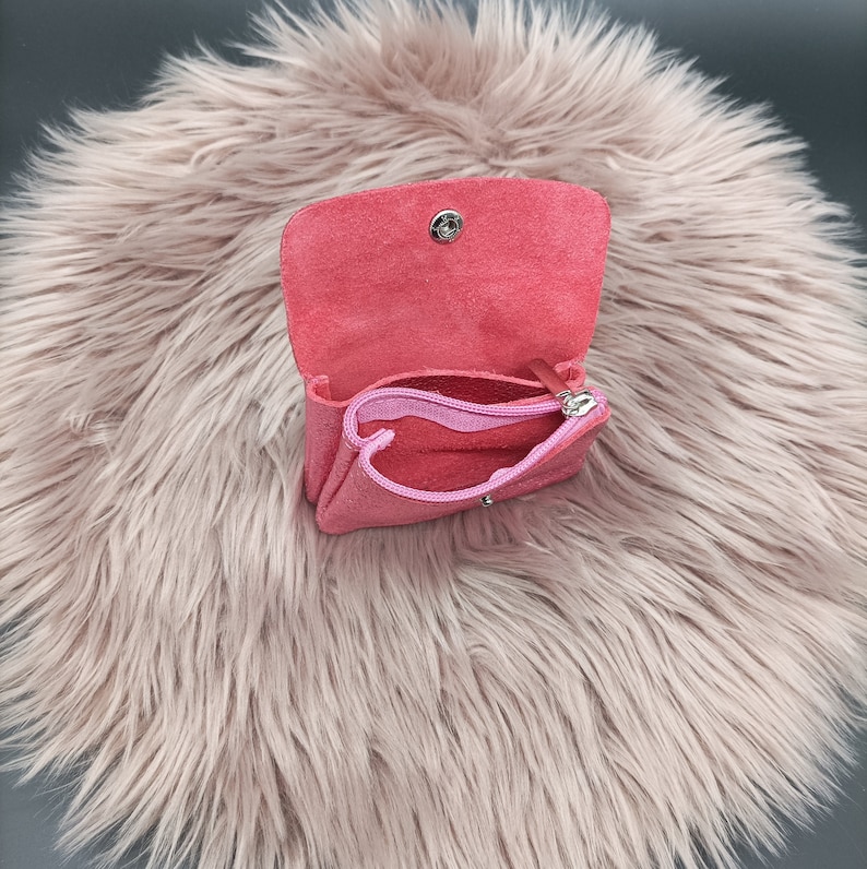May include: A small, open, pink leather coin purse with a matching pink zipper pouch inside. The purse has a snap closure and is set against a fluffy, light pink background.
