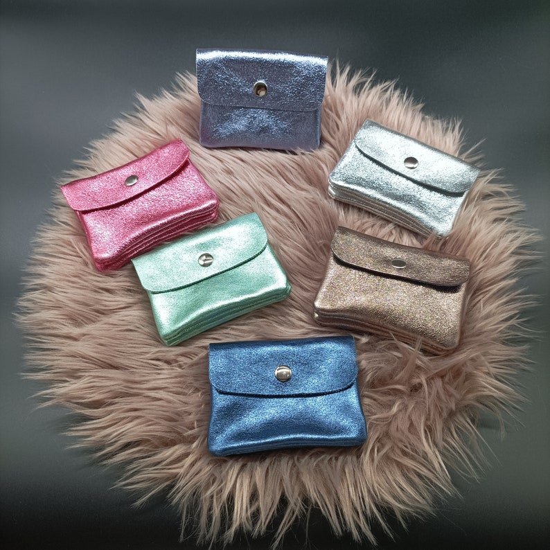 May include: A collection of small, glittery coin purses in various colors, including pink, blue, silver, and gold. Each purse has a flap closure with a silver snap. The purses are arranged on a fluffy, pink surface.