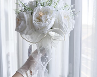 May include: A white bridal bouquet featuring large, open roses and delicate lily of the valley. The bouquet is accented with a sheer, white ribbon adorned with pearls and held by a white gloved hand.