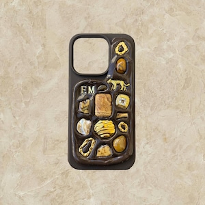 May include: A brown phone case with a textured surface and gold accents. The case has a variety of small, decorative elements, including a small animal figure, a ring, and several stones. The initials "EM" are embossed on the case.