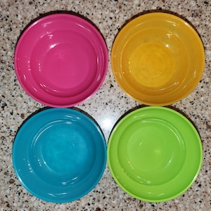May include: Four colorful plastic bowls in vibrant shades of pink, yellow, teal, and green. The bowls are round with a slightly raised rim, perfect for serving snacks or small portions.