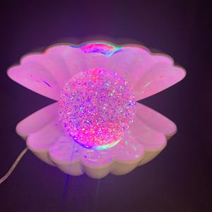 May include: A seashell-shaped lamp with a glittery sphere at its core. The lamp emits a soft, colourful light, creating a whimsical ambiance. The shell is white with pink and purple hues. The lamp is powered by a white cable.