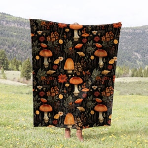 May include: A black throw blanket with a repeating pattern of orange and brown mushrooms and flowers. The blanket is being held up in front of a green grassy field and a mountain range in the background.