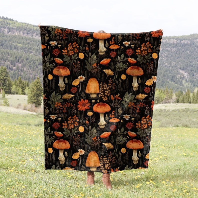 May include: A black throw blanket with a repeating pattern of orange and brown mushrooms and flowers. The blanket is being held up in front of a green grassy field and a mountain range in the background.