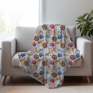 May include: A blue and white throw blanket with a baseball themed pattern. The pattern includes baseballs, bats, gloves, hot dogs, and popcorn.