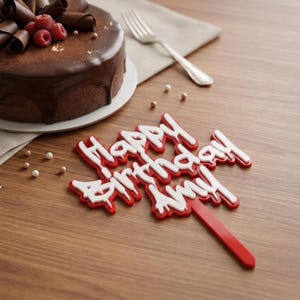 May include: A chocolate cake with chocolate shavings and fresh raspberries sits next to a fork and a cake topper that says "Happy Birthday Amy" in red and white lettering. The cake is on a white plate.