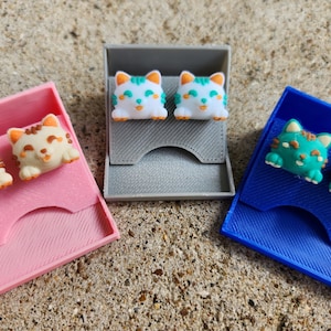 Könnte beinhalten: Drei kleine Boxen mit unterschiedlich farbigen Deckeln, jede mit einem Paar katzenförmiger Ohrringe. Die Ohrringe sind aus weißem, hellblauem und grünem Material.