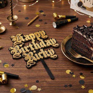 May include: A gold and black "Happy Birthday Ricky" cake topper with a slice of chocolate cake on a black plate. The cake is topped with cookie pieces. Party horns, confetti, and a candle holder are also visible.