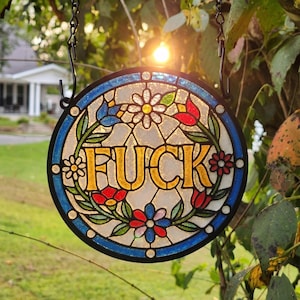 May include: A circular stained glass suncatcher featuring the word FUCK in bold yellow letters at the center. The design is surrounded by a floral pattern with red, blue, and green glass pieces, all enclosed within a black metal frame and chain.