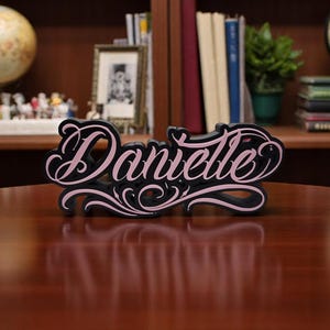 Puede incluir: Una placa de nombre de madera negra con el nombre "Danielle" en una fuente cursiva, delineada en rosa. La placa tiene remolinos decorativos y se muestra sobre una mesa de madera.